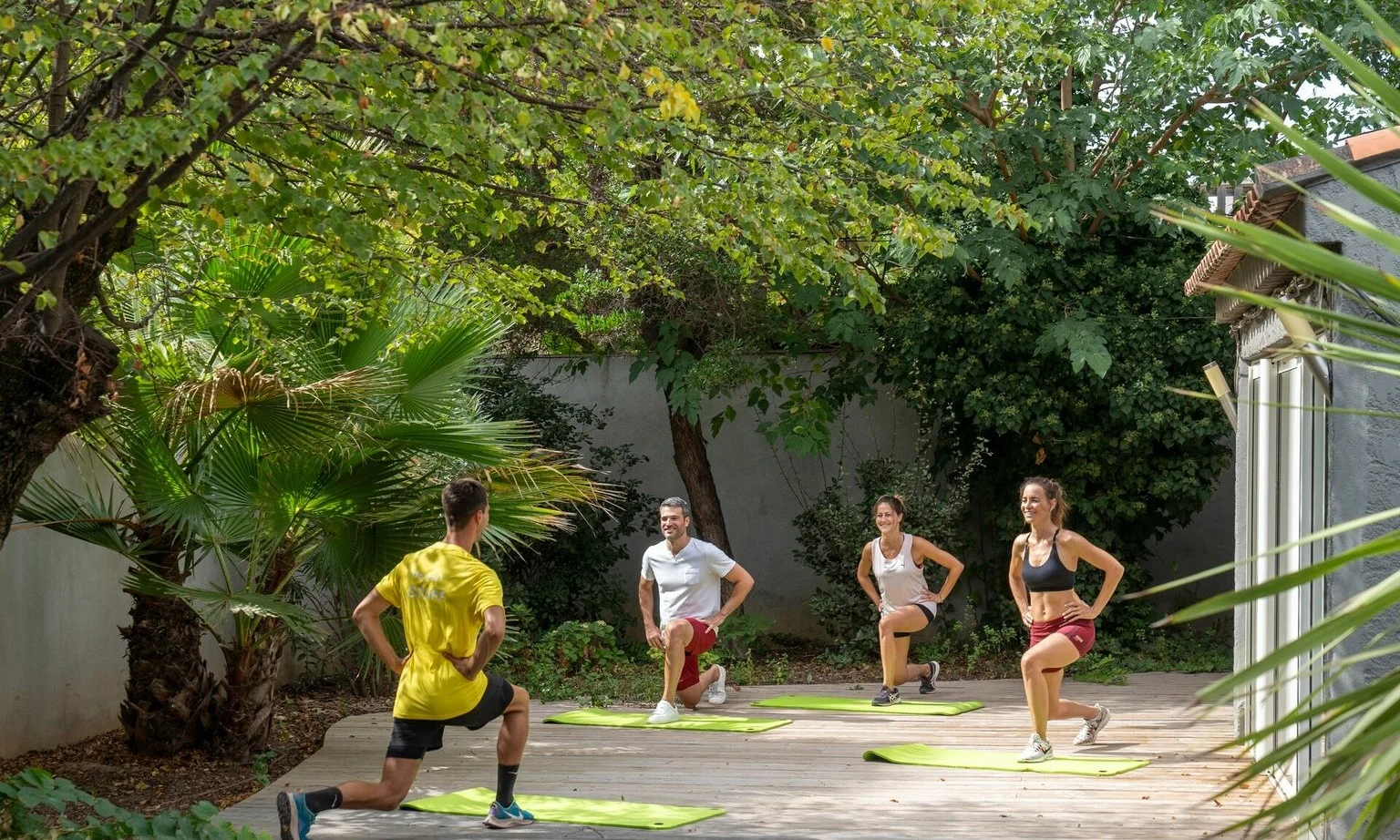 Four people exercising lunges on yoga mats, under lush green trees near a building at La Barque