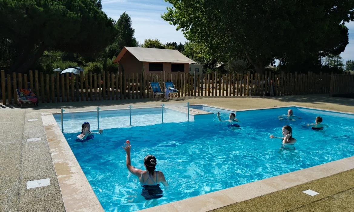 People swimming in a clear blue pool surrounded by trees and wooden fencing, with tents and lounge chairs nearby at Seasonova Ile de Ré