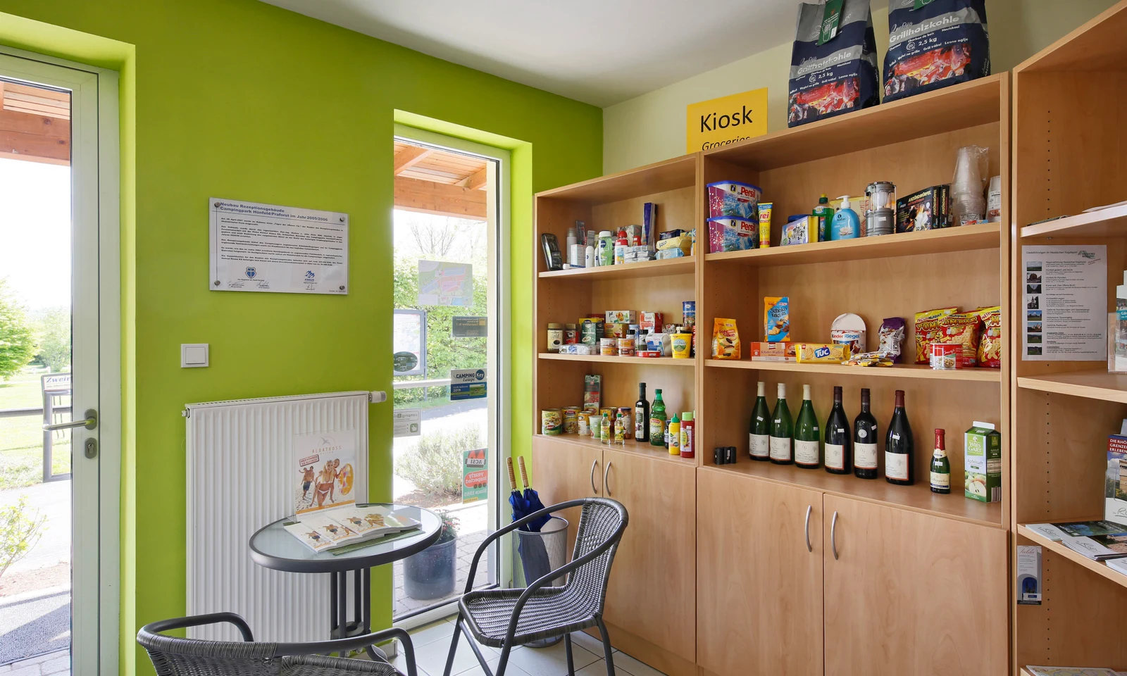 Shelves holding assorted groceries and drinks, with a small table and chairs, inside a bright room at KNAUS Campingpark Hünfeld