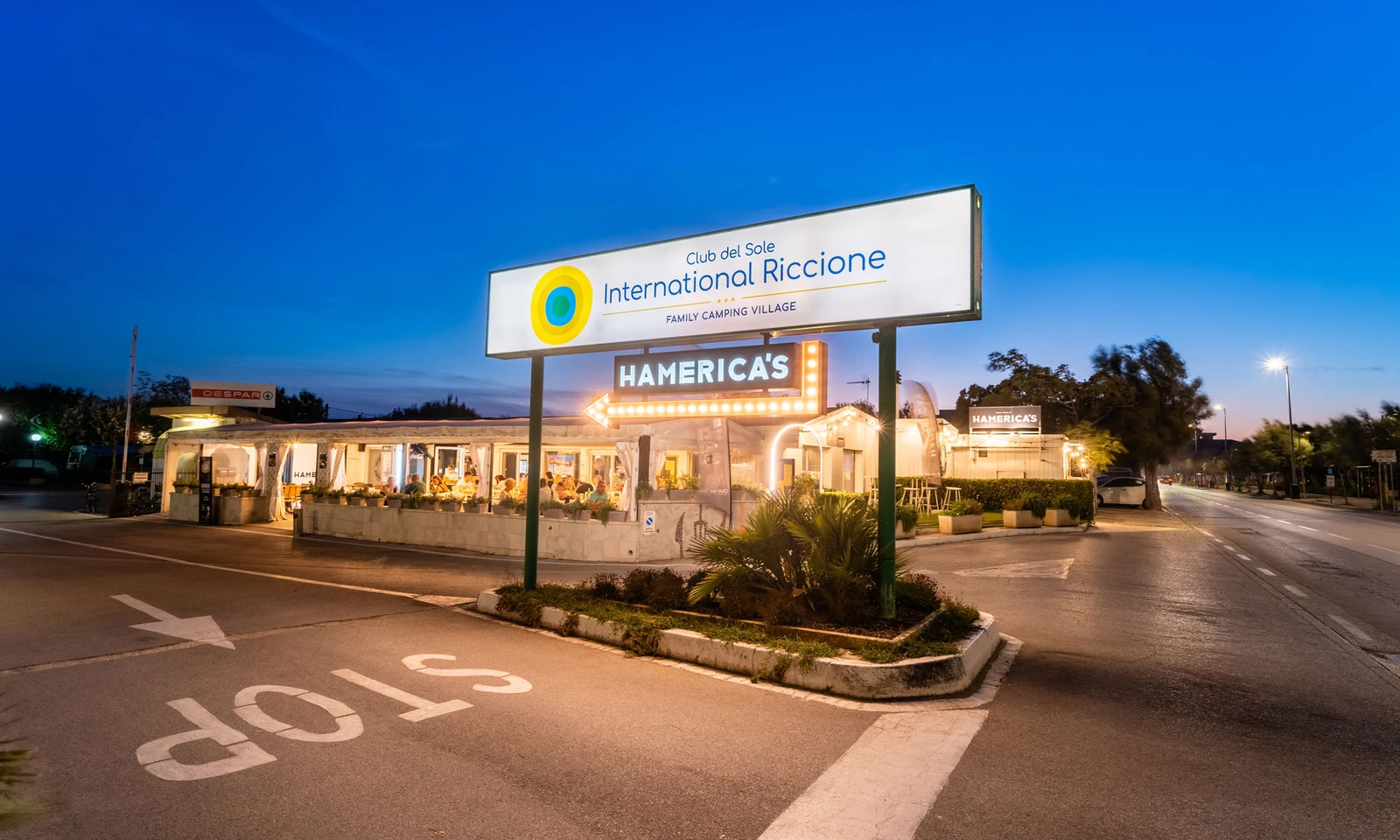 Restaurant with illuminated signs, bustling with people dining outdoors, located beside a road during evening hours at International Riccione Family Camping Village