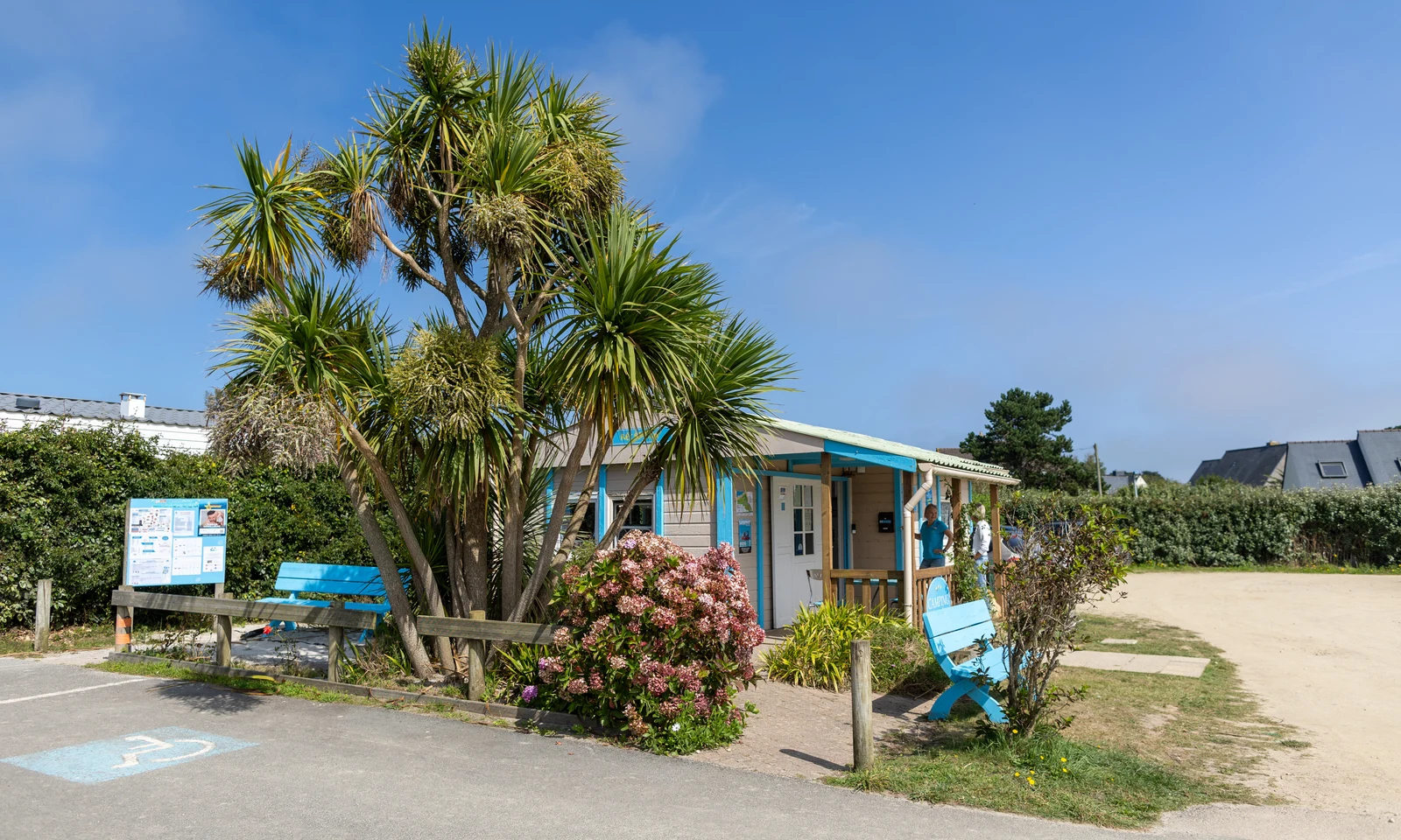 Small wooden building with people standing outside, surrounded by lush greenery and blue benches, under clear skies at La Pointe de Roscoff