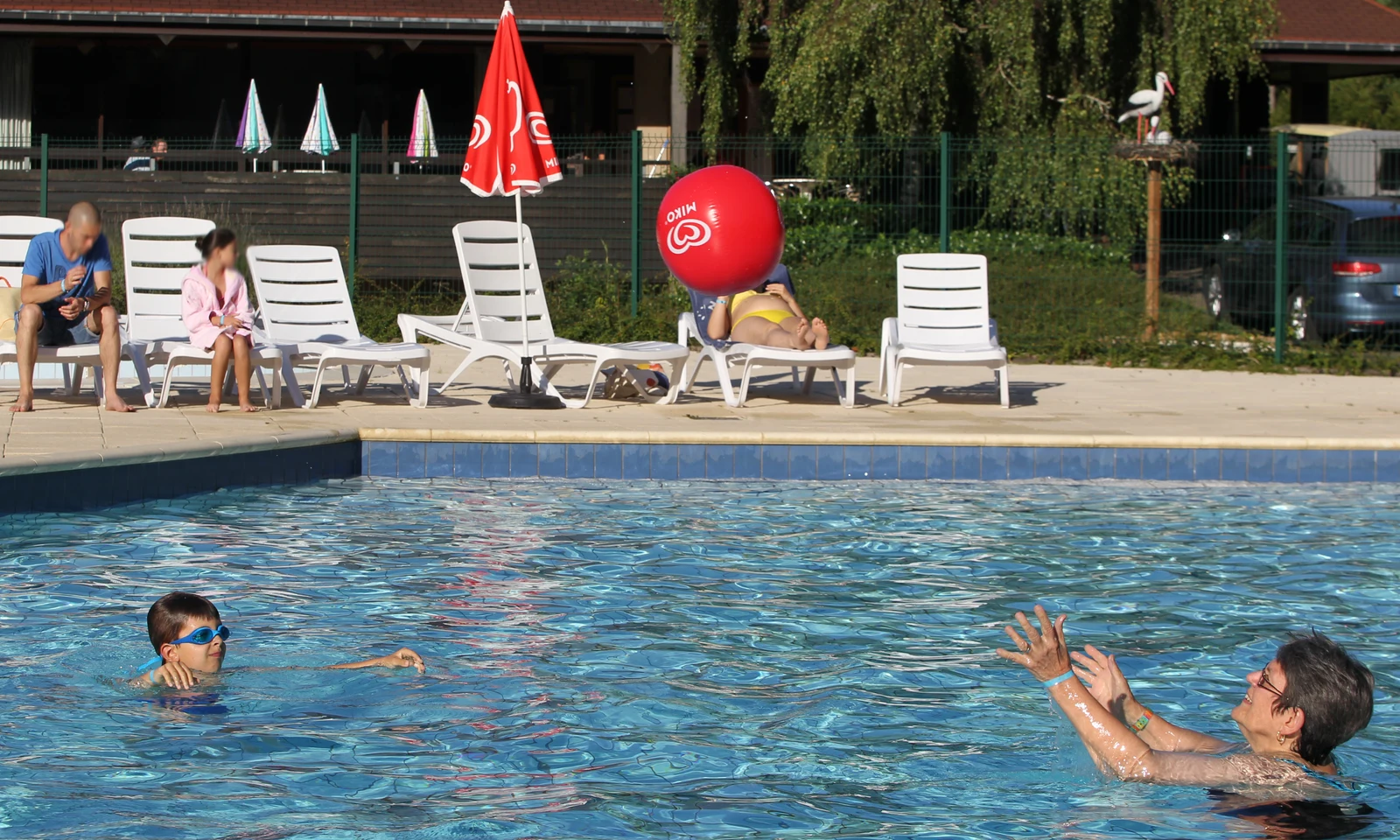 Child and adult playing in a swimming pool; people relaxing on deck chairs nearby under sunny skies at La Foret