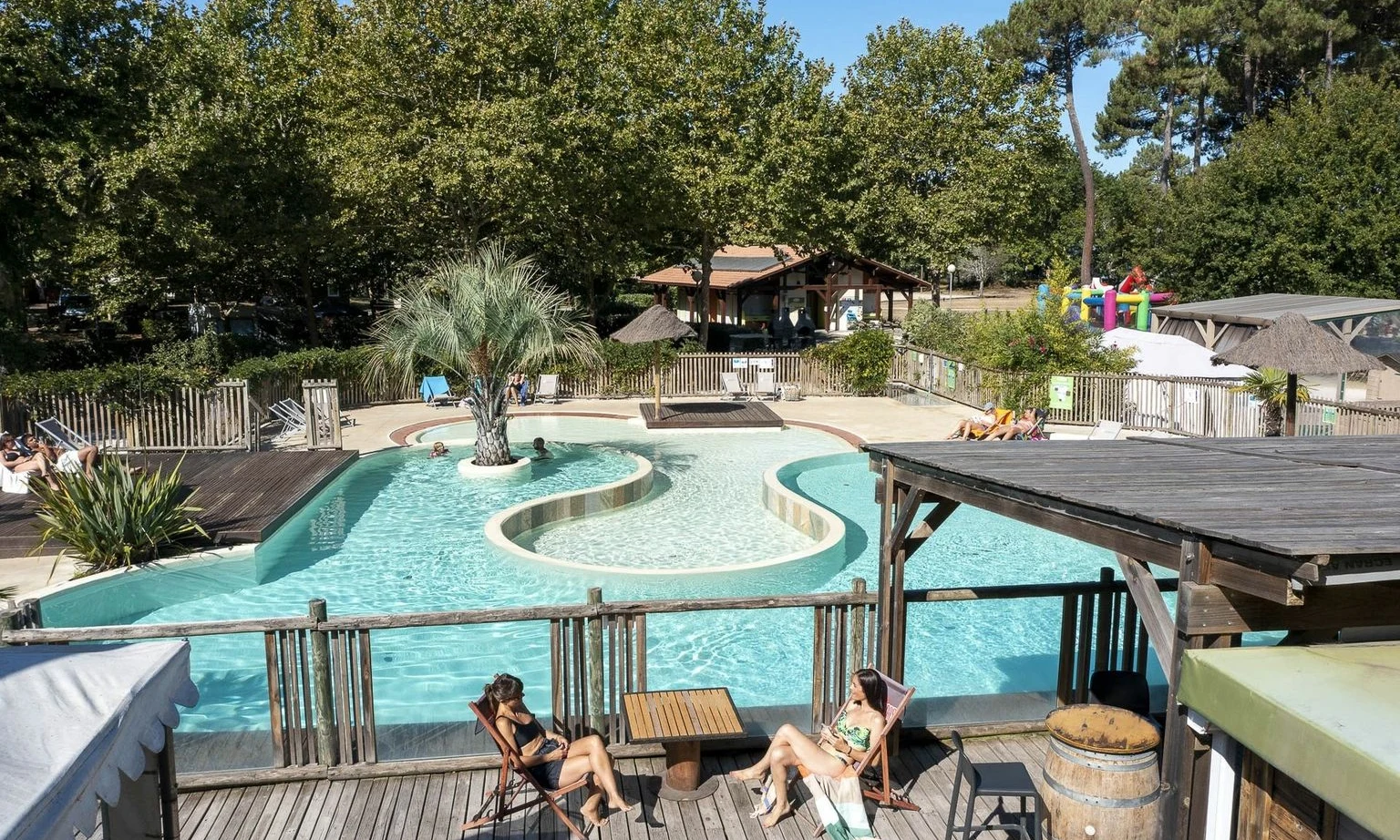 Two women relax on lounge chairs beside a pool surrounded by trees and a wooden deck at Lac de Sanguinet