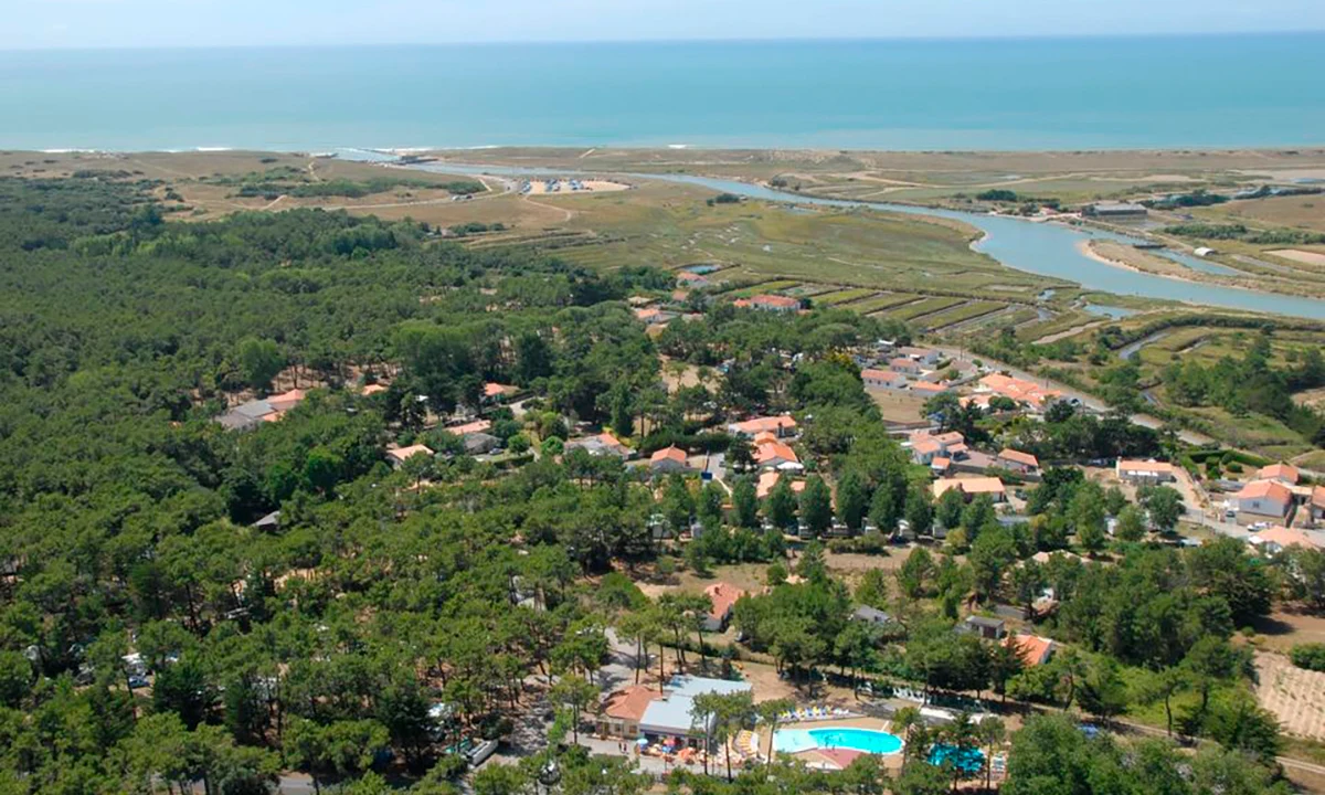 Buildings nestled amidst dense trees near a small lake, with a vast coastline and ocean visible in the distance at Camping 2 Plages & Océan Village Corsaire