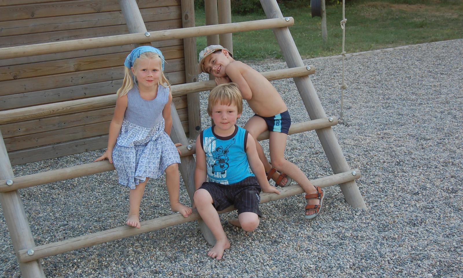 Three children sitting and leaning on a wooden climbing structure, in a gravel playground area at Les Arches