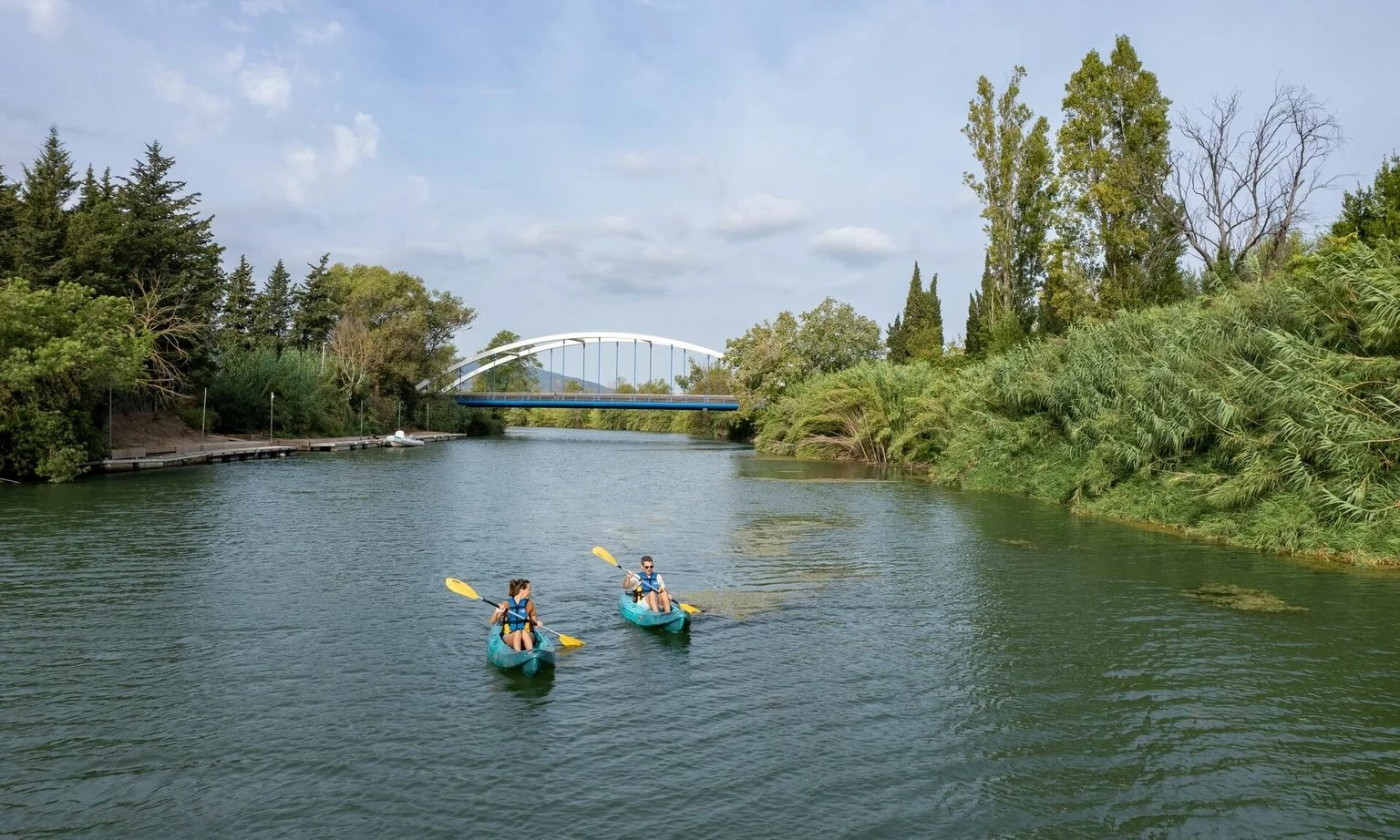 Two people kayaking on a river, surrounded by dense greenery with a bridge in the background at La Barque