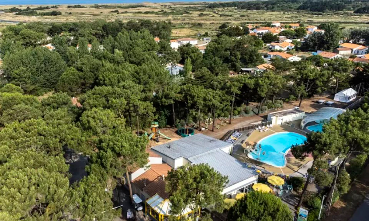 Aerial view of a swimming pool surrounded by buildings and dense trees, with rural landscape in the background at Camping 2 Plages & Océan Village Corsaire