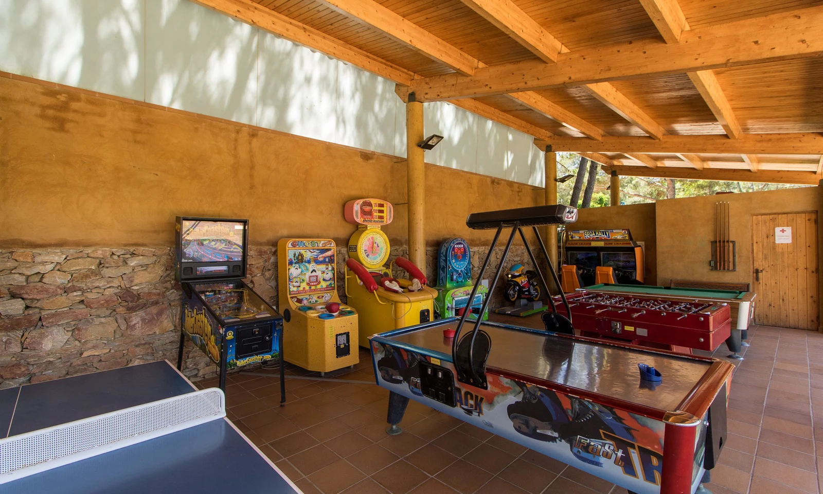 Arcade games and tables are arrayed in a covered recreation area with wooden beams and stone walls at Begur