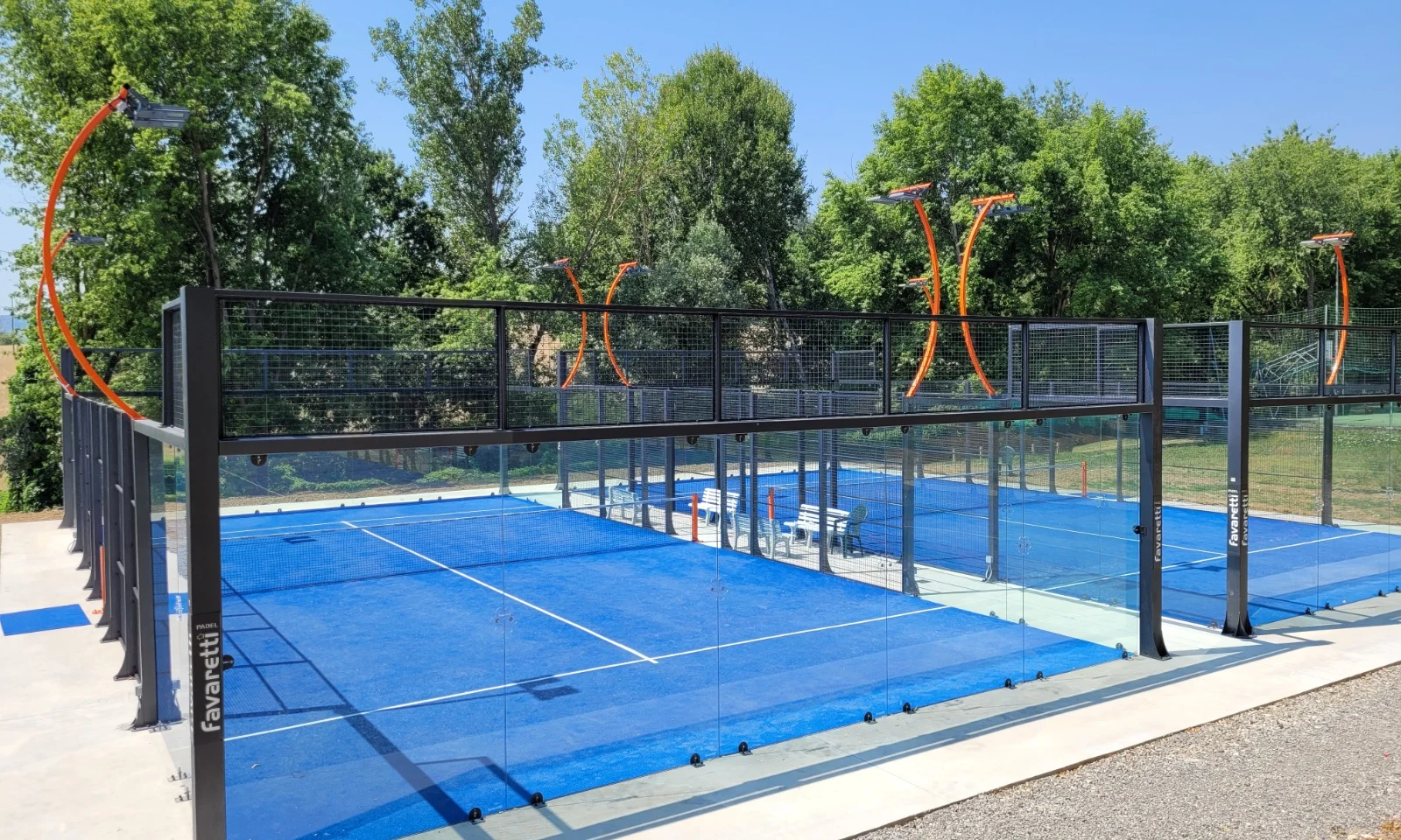 Blue padel courts surrounded by glass walls, sunlight, and green trees in the background at Isamar