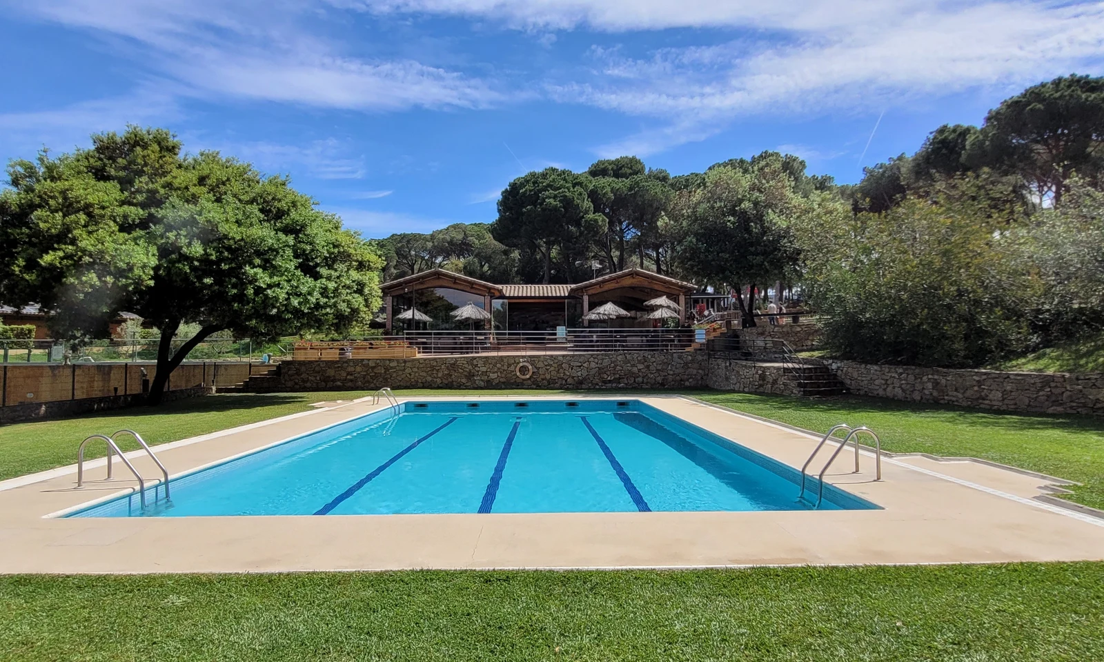 Swimming pool with ladders in lush outdoor setting near stone buildings and shaded picnic area at Begur