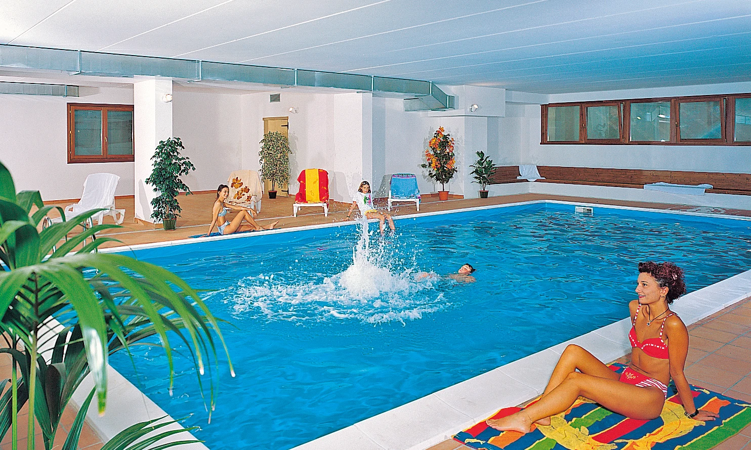 People enjoying swimming and relaxing around an indoor pool with plants, lounge chairs, and windows at Residence Campi