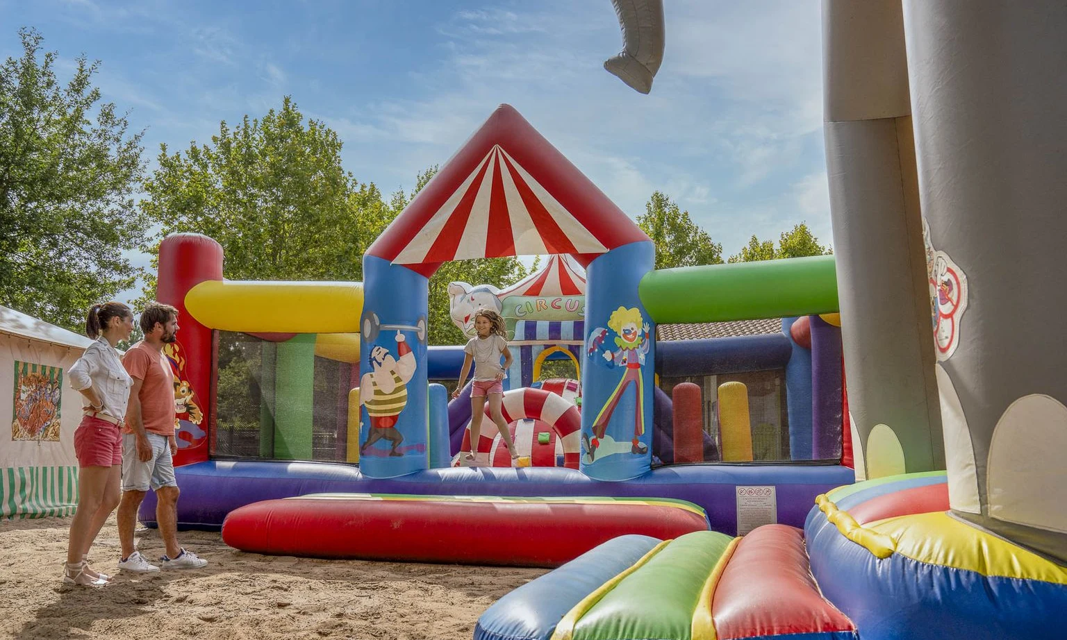 Inflatable bouncy castle with kids playing, observed by adults, surrounded by trees and a tent at Lac de Sanguinet