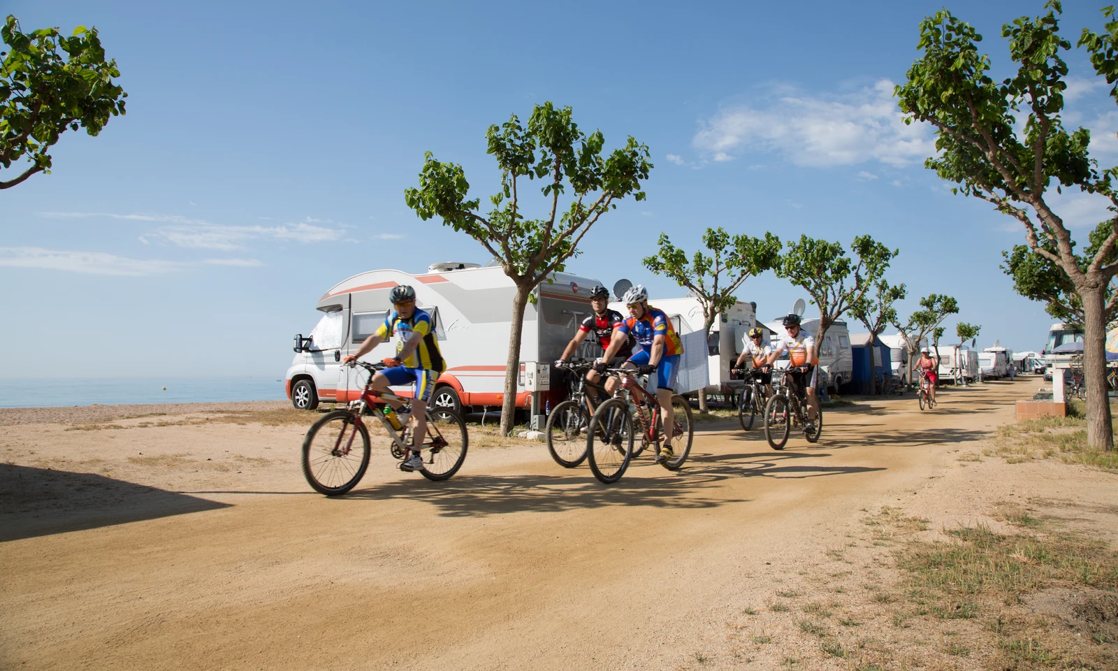 Cyclists riding on a dirt path, passing parked caravans and trees near the beach at Bon Repos