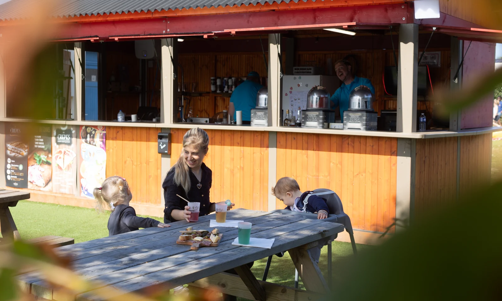 A woman and two children sit at a picnic table eating snacks near an outdoor food stall at La Pointe de Roscoff