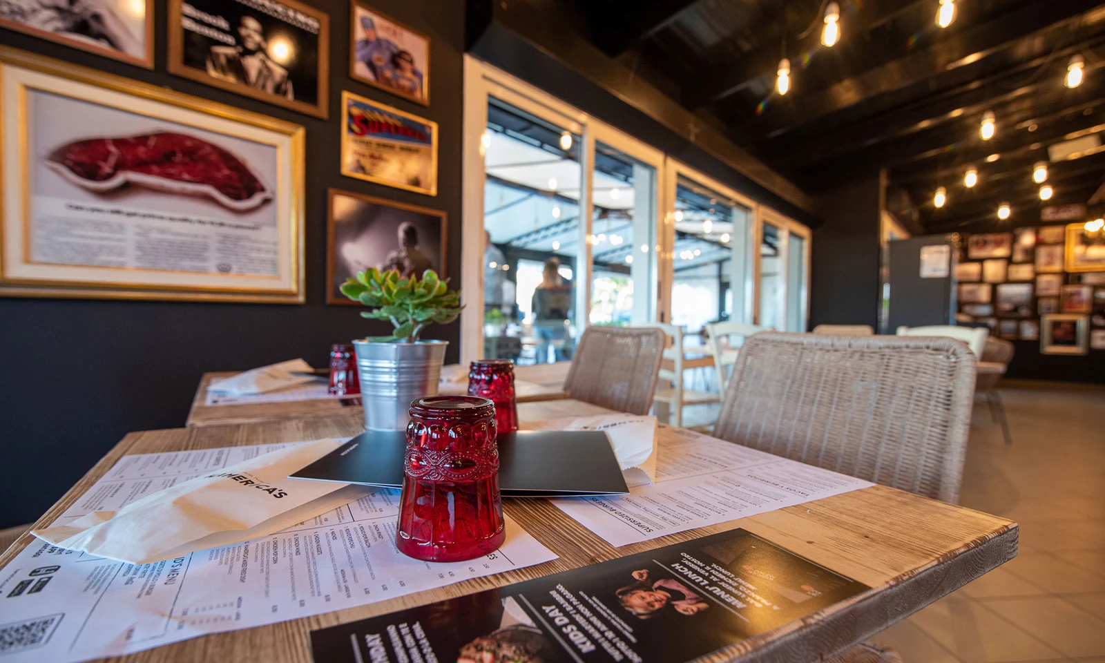 Red candle holders and a potted plant decorate a wooden restaurant table, surrounded by framed pictures on walls at International Riccione Family Camping Village