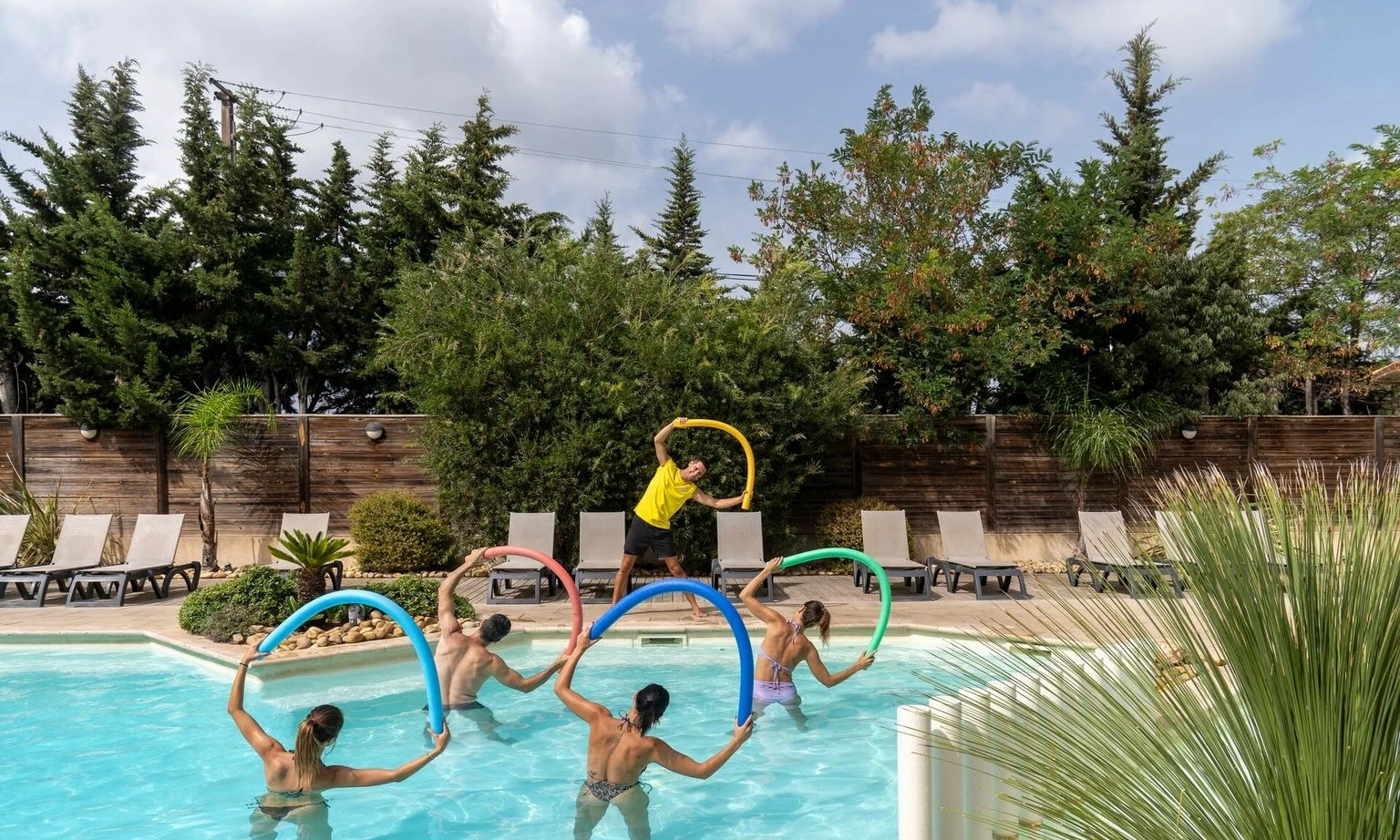 People participating in a pool exercise class using pool noodles, surrounded by lounge chairs and lush greenery at La Barque