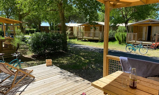 Wooden deck with chairs and table, shaded by a roof, overlooking nearby cabins and bicycles under trees at Seasonova Ile de Ré