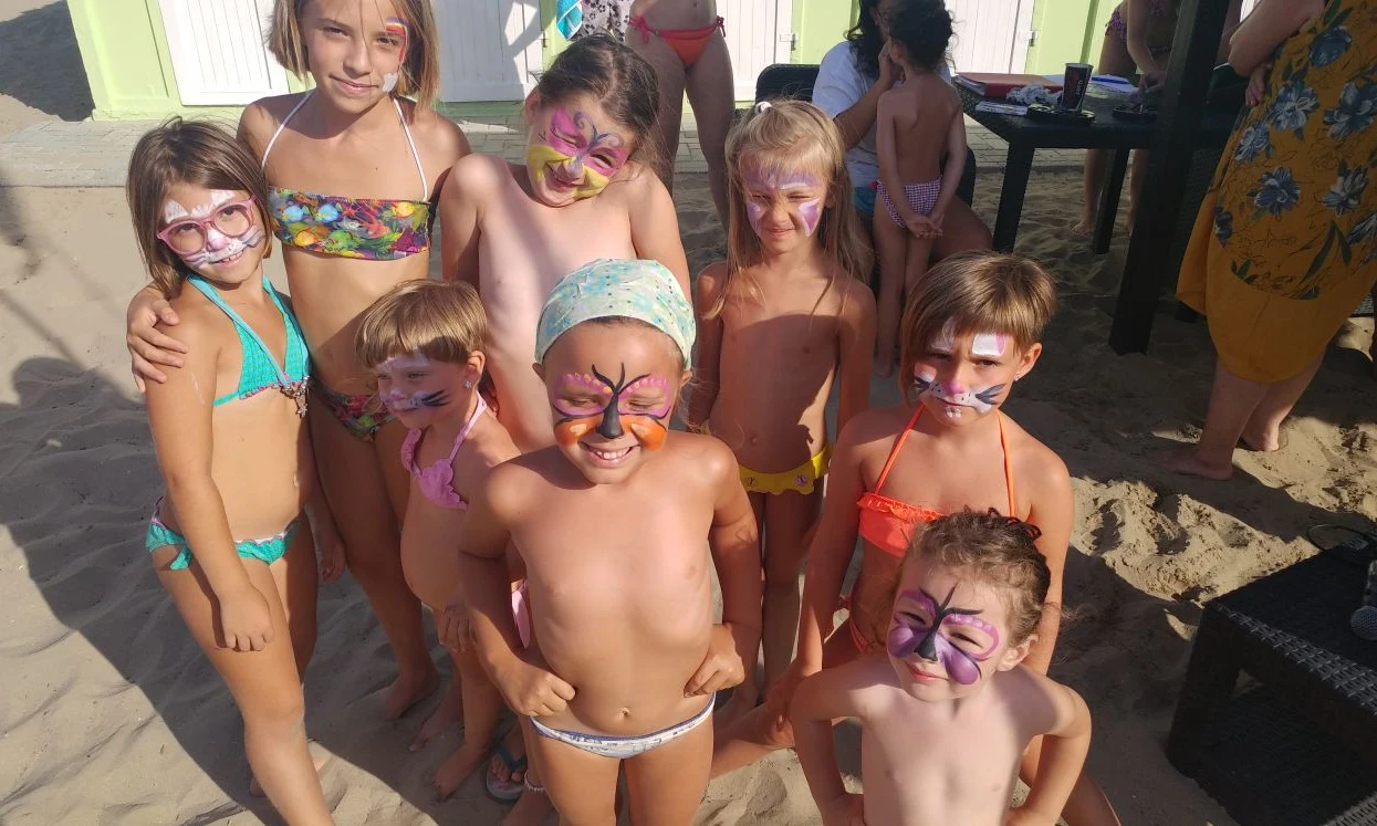 Children with painted faces smile, standing on sandy ground, wearing swimsuits under the sun at International Riccione Family Camping Village