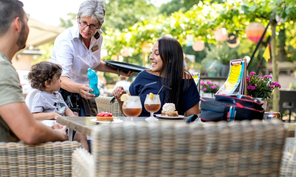 Waitress serves drinks to a smiling family at an outdoor cafe with lush greenery and hanging lanterns at Landal Rabbit Hill