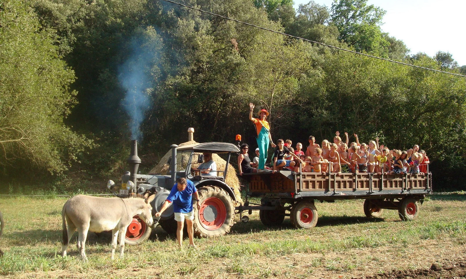 A tractor pulling a trailer filled with people waving, with a man leading a donkey, in a rural setting at Begur