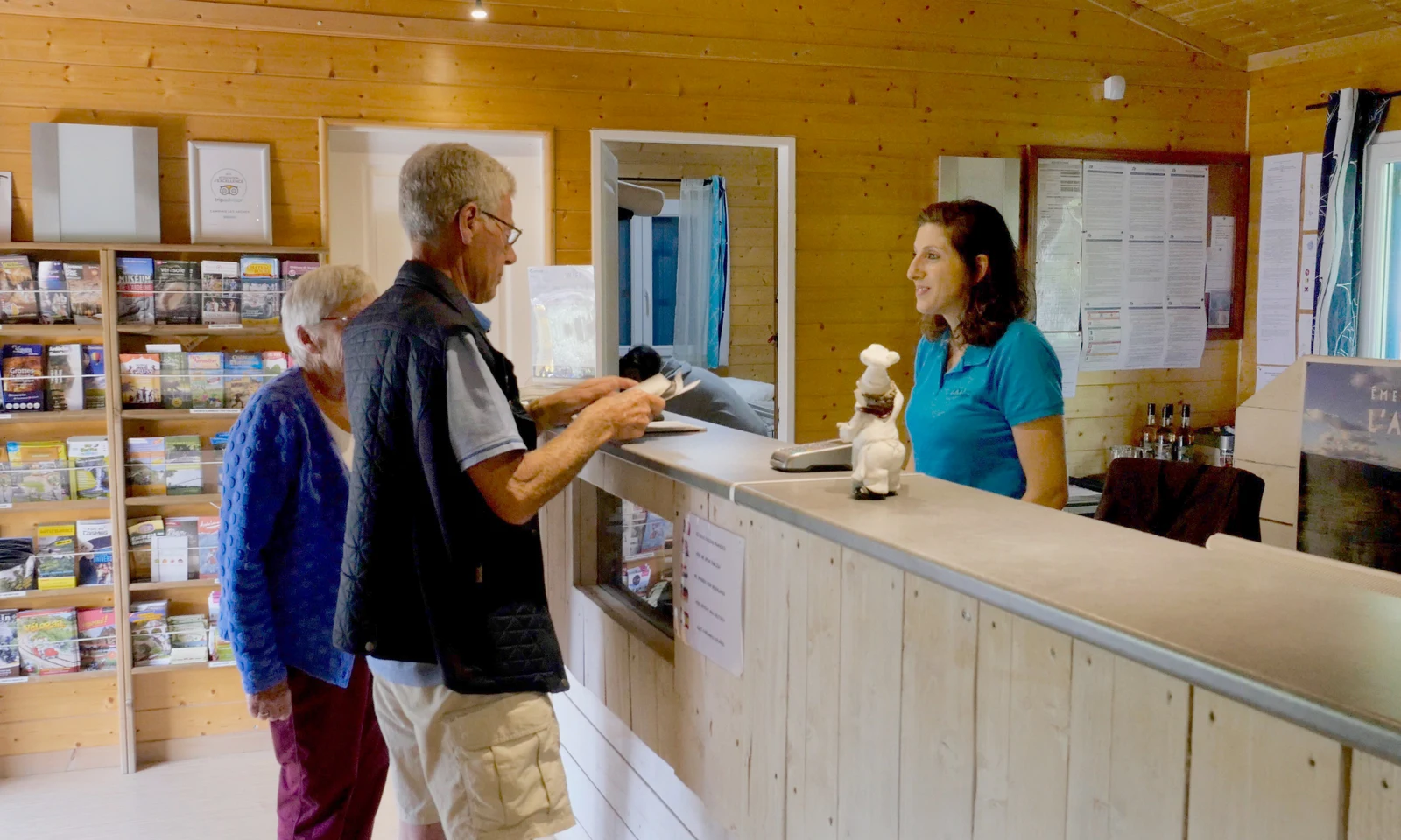 Two individuals inquire at a reception desk with a staff member in a wooden-paneled information office at Les Arches