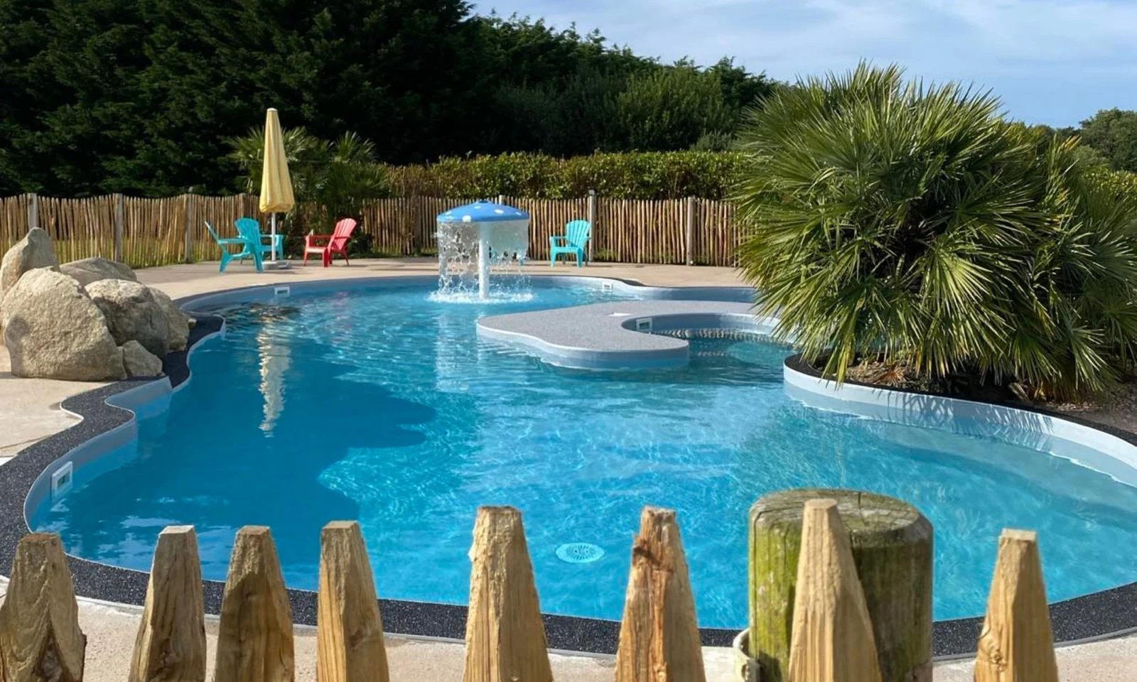 A small pool with water fountain surrounded by palm trees and colorful chairs, enclosed by wooden fence at Domaine de Mesqueau