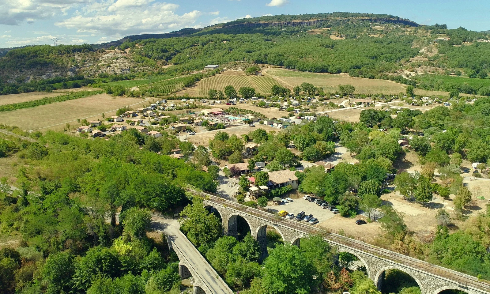 Railway bridge spans valley amidst a campsite with cabins and greenery, surrounded by rolling hills and farmland at Les Arches