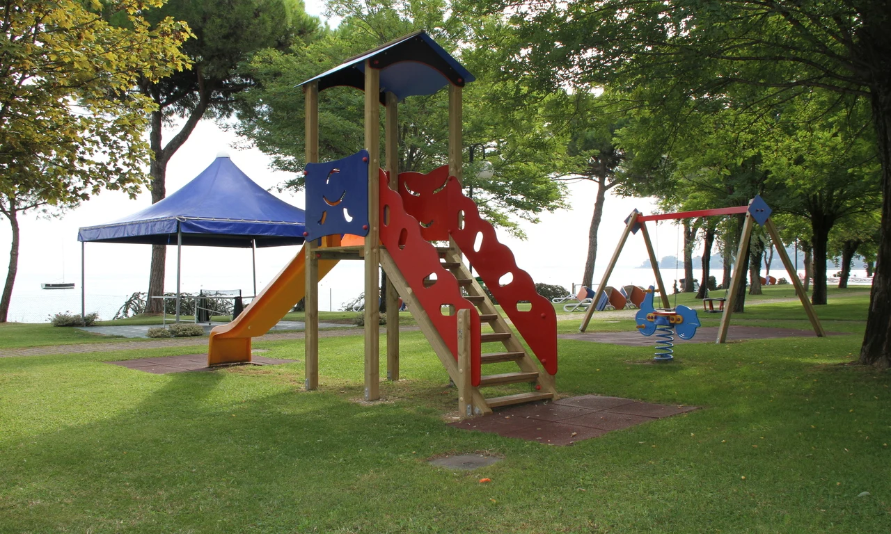 Playground set with slides and swings on grass under trees near a tent, adjacent to a lakeside view at Front Lake Resort Le Corti del Lago