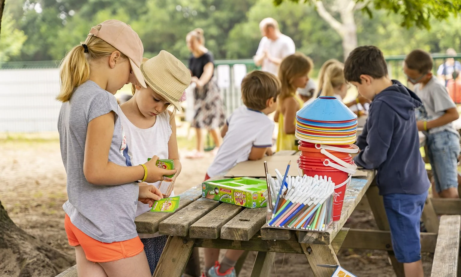 Children engaging in arts and crafts around a picnic table, surrounded by trees and other children in the background at Lac de Sanguinet