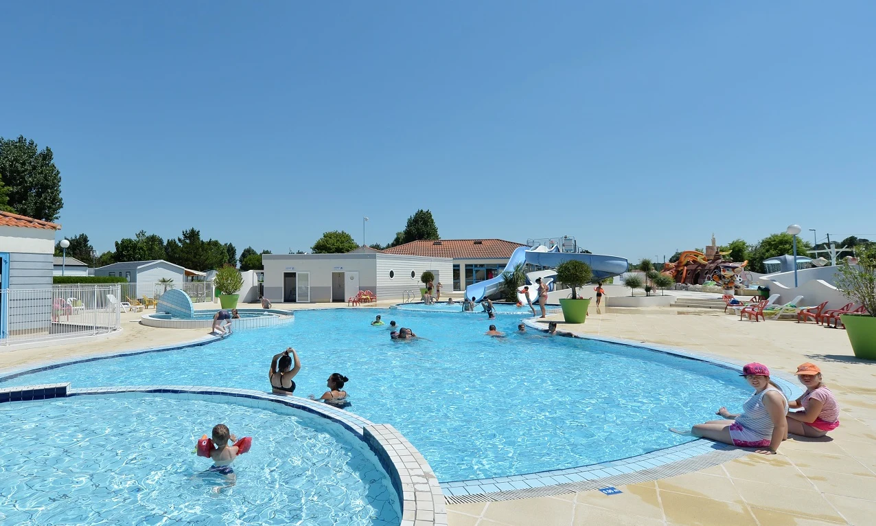 People swim and relax in a large pool near buildings and waterslides, on a sunny day at Les Aventuriers de la Calypso