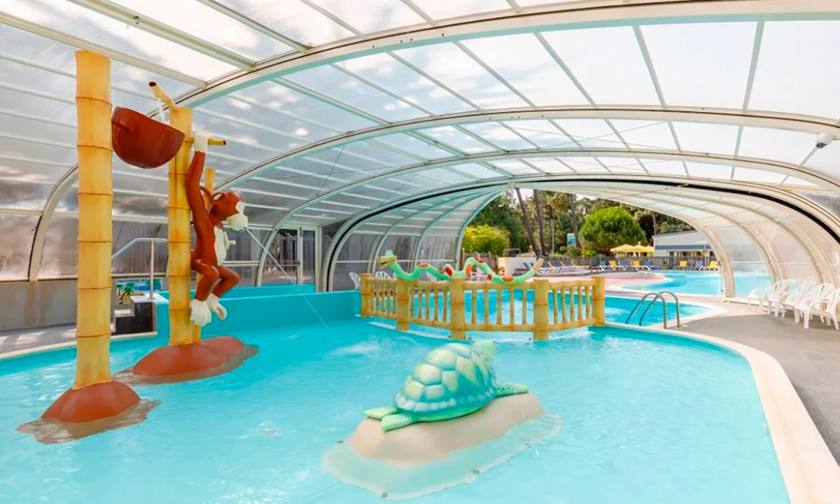 Indoor pool with water fountains and animal-themed structures, including a monkey on a tree, under a clear cover at Camping 2 Plages & Océan Village Corsaire