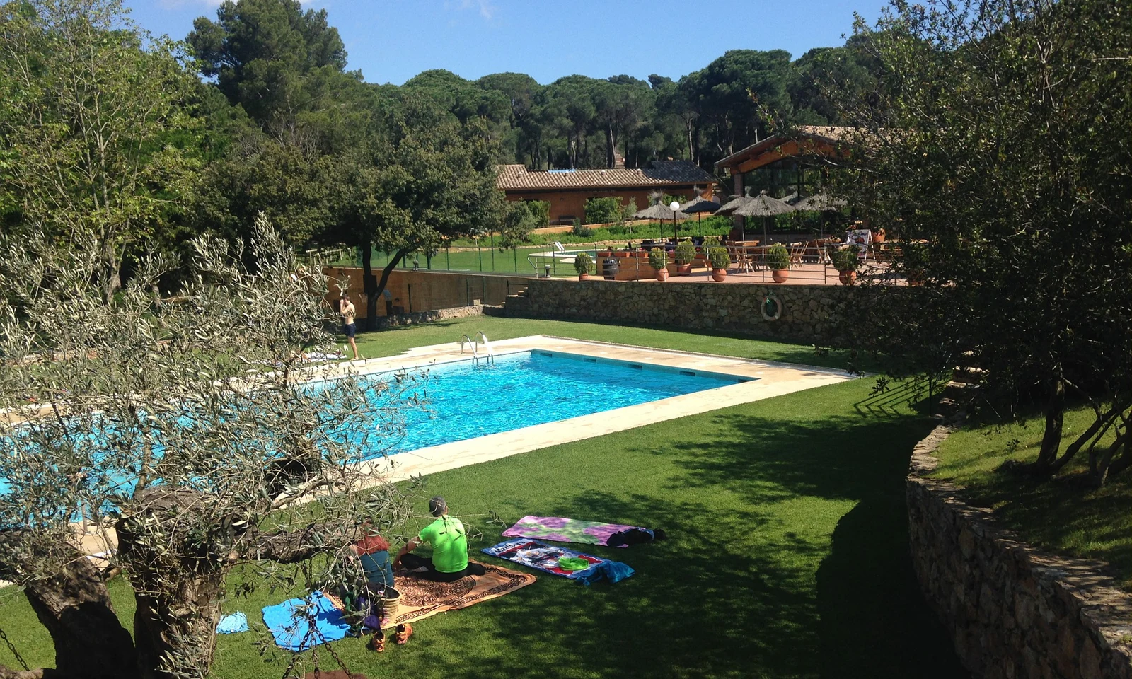 Swimming pool surrounded by trees with people sunbathing on the grass, near a stone patio with outdoor seating at Begur