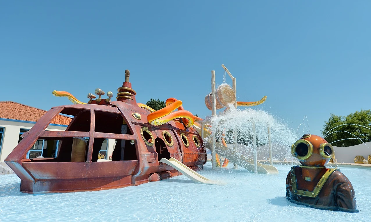 Colorful submarine-themed water play structure splashing water in shallow pool, complemented by a deep-sea diver statue at Les Aventuriers de la Calypso