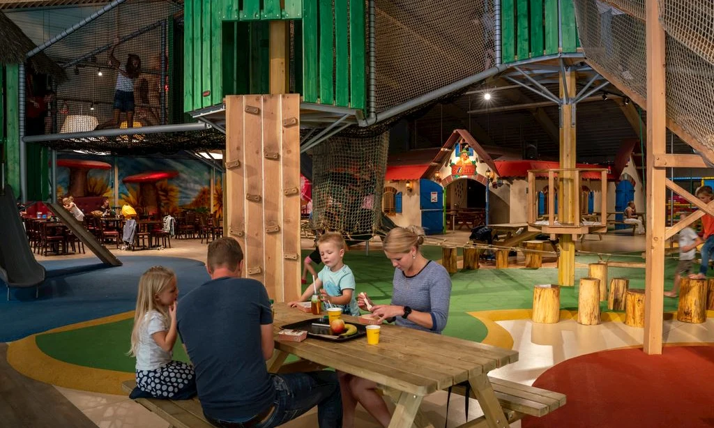 Family eating at a wooden table while children play on indoor climbing structures and slides at Landal Rabbit Hill