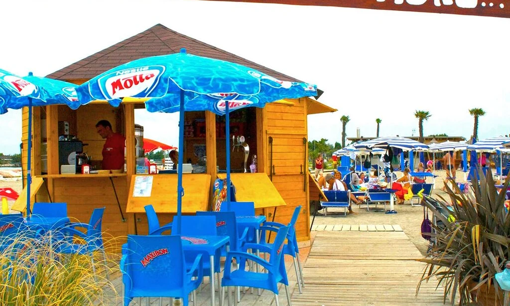 Wooden beach kiosk serving, surrounded by blue chairs and umbrellas, with people relaxing on the sandy beach at Oasi