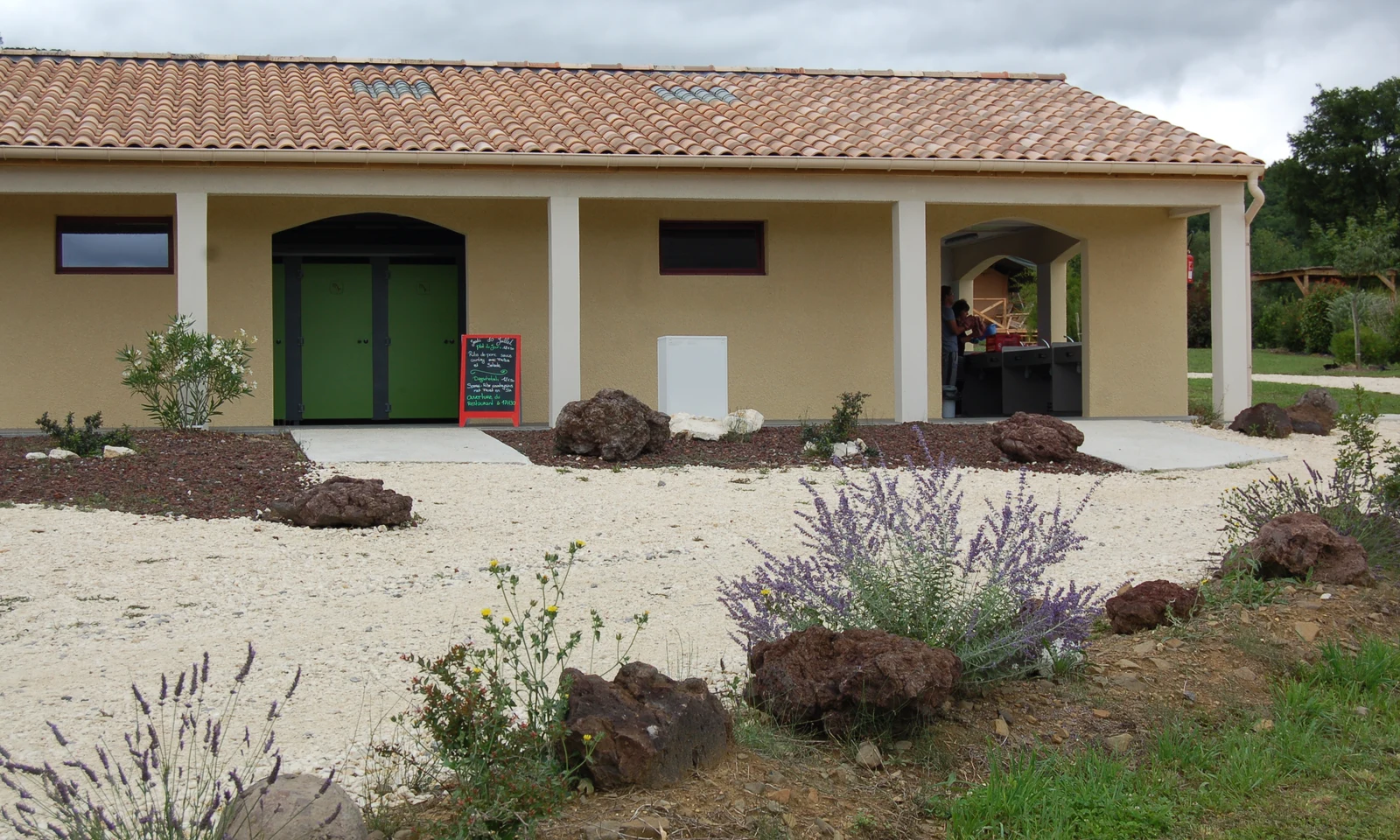 Building with green doors, surrounded by plants and rocks, housing sinks under a covered area at Les Arches