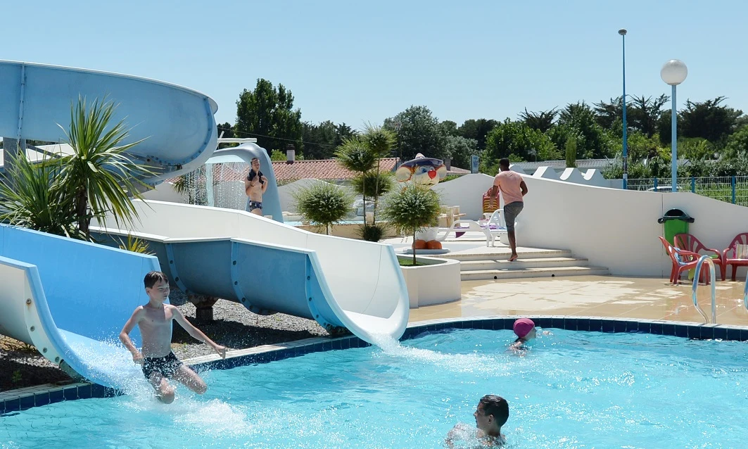 Child sliding into a swimming pool, another child swimming, and people relaxing around at Les Aventuriers de la Calypso