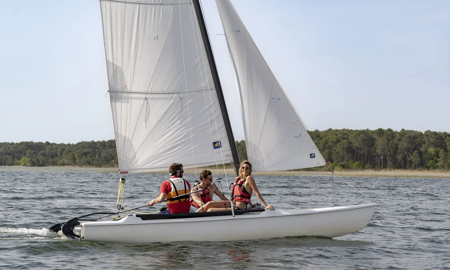 Sailboat carrying three people gliding on a lake with a forest shore in the background at Lac de Sanguinet