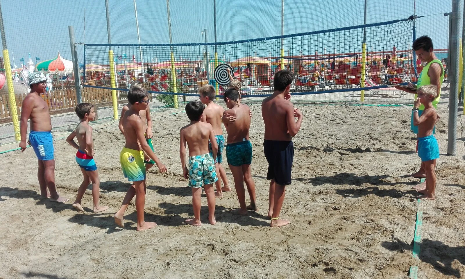 Children stand attentively in a sandy area, listening to an instructor near a net at International Riccione Family Camping Village