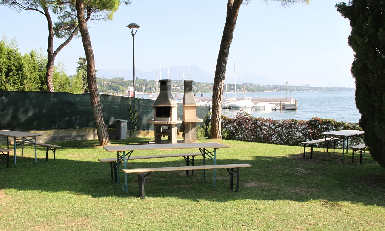 Barbecue grills stand unused between picnic tables, trees, and a lakeside with docked sailboats in the background at Front Lake Resort Le Corti del Lago