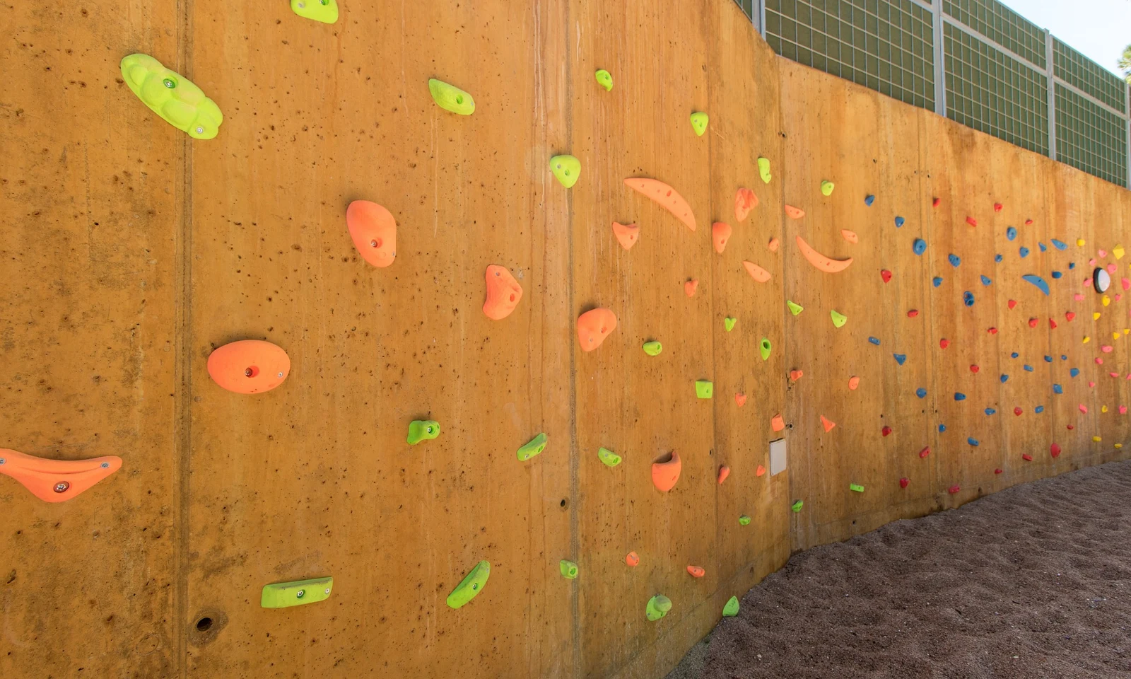 Climbing wall with colorful handholds, set against a curved, yellow concrete surface, surrounded by a gravel area at Begur