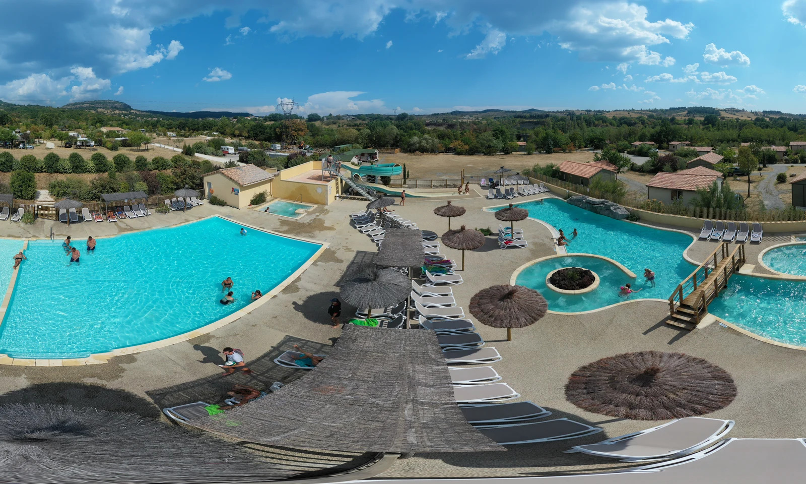 Two swimming pools with people swimming and sunbathing surrounded by loungers, thatched umbrellas, and greenery at Les Arches