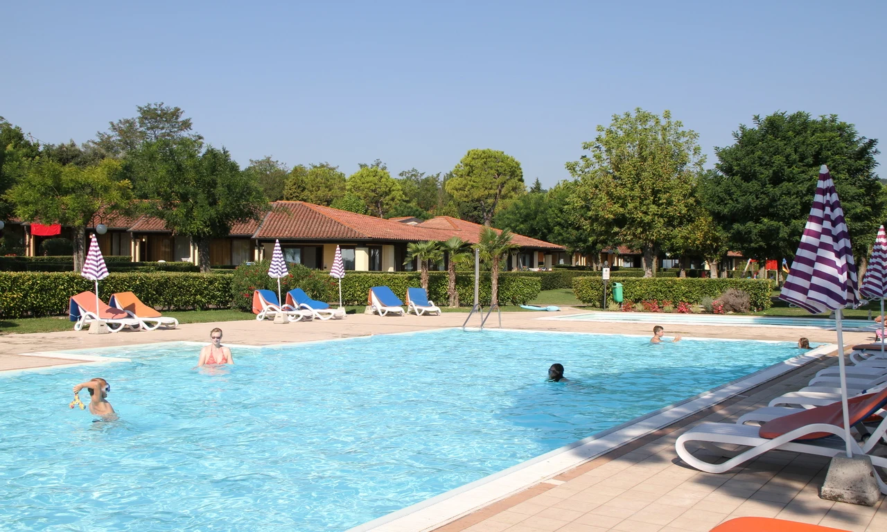 Swimming pool occupied by people, sunbeds and umbrellas surrounding it, with bungalows and lush green trees in the background at Front Lake Resort Le Corti del Lago