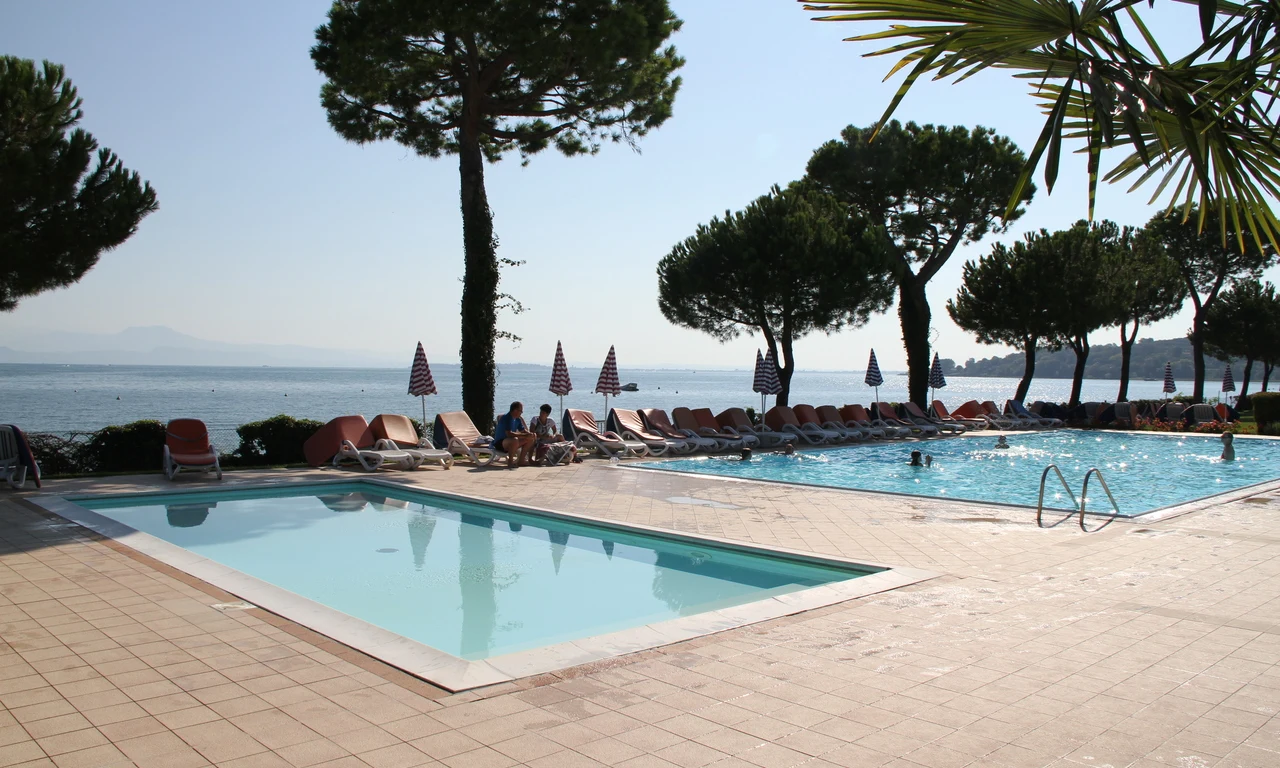 Two swimming pools with people relaxing on lounge chairs, surrounded by trees, overlooking a serene lake at Front Lake Resort Le Corti del Lago