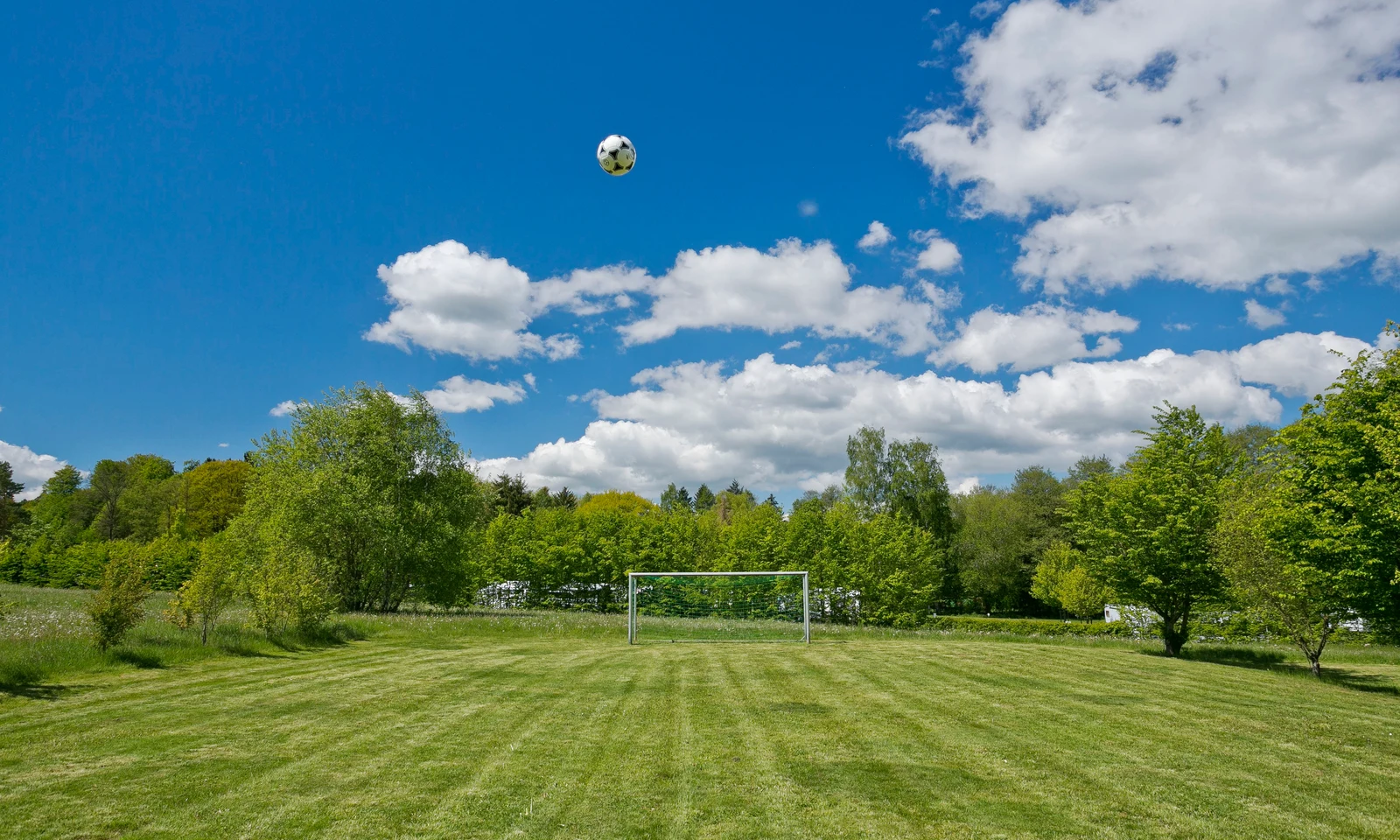 Soccer ball flying above a goalpost on a grassy field, surrounded by trees and under a clear blue sky at KNAUS Campingpark Hünfeld