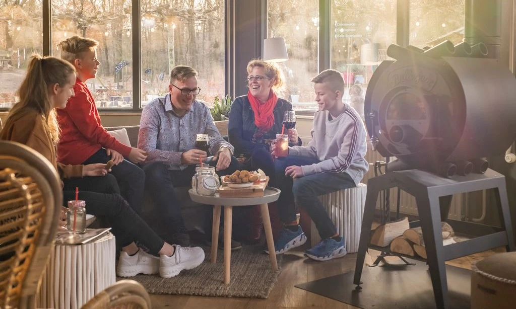 Five people sit on a sofa and chairs, sharing drinks and snacks in a cozy, sunlit room at Landal Rabbit Hill