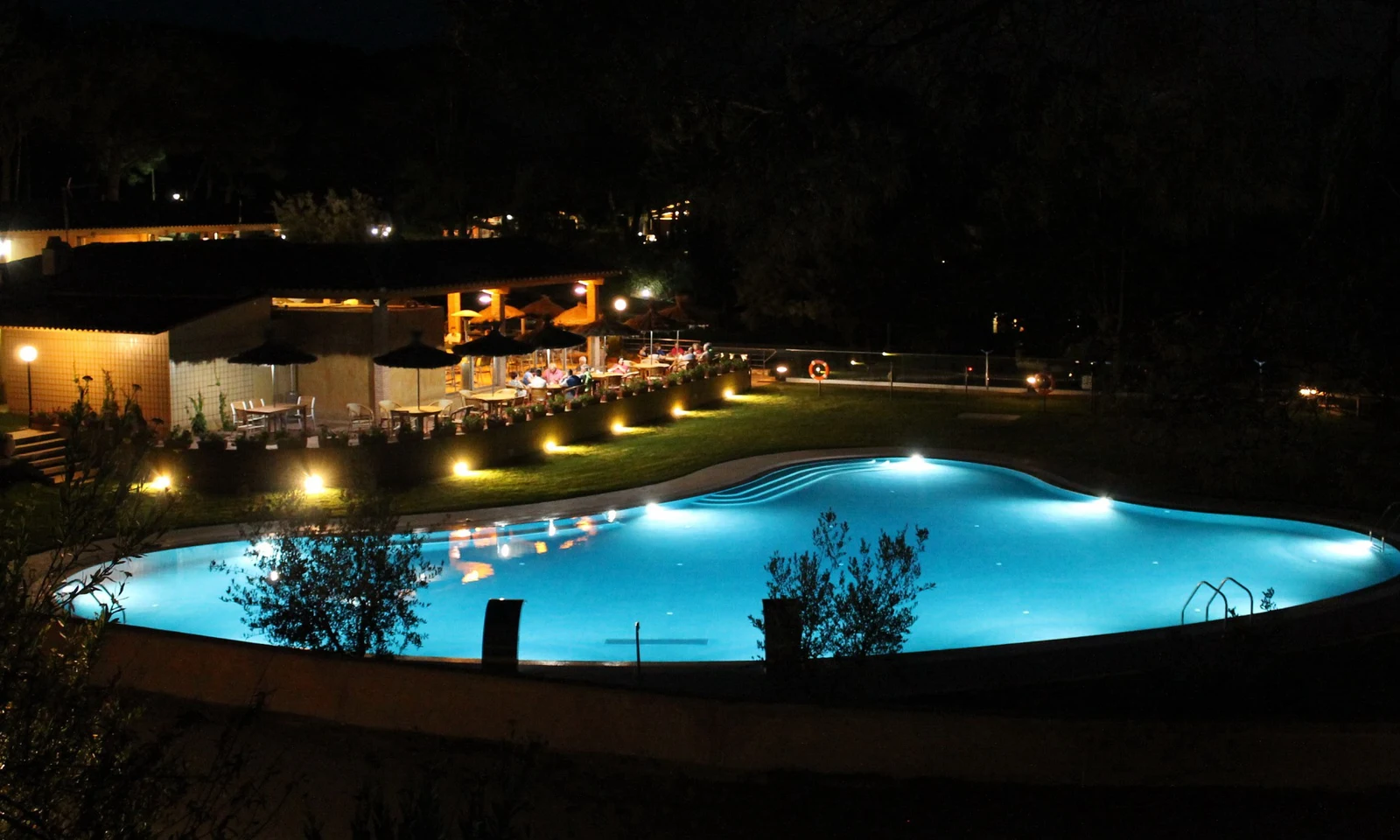 Illuminated swimming pool glowing at night, surrounded by a grassy area and a restaurant with people dining outdoors at Begur