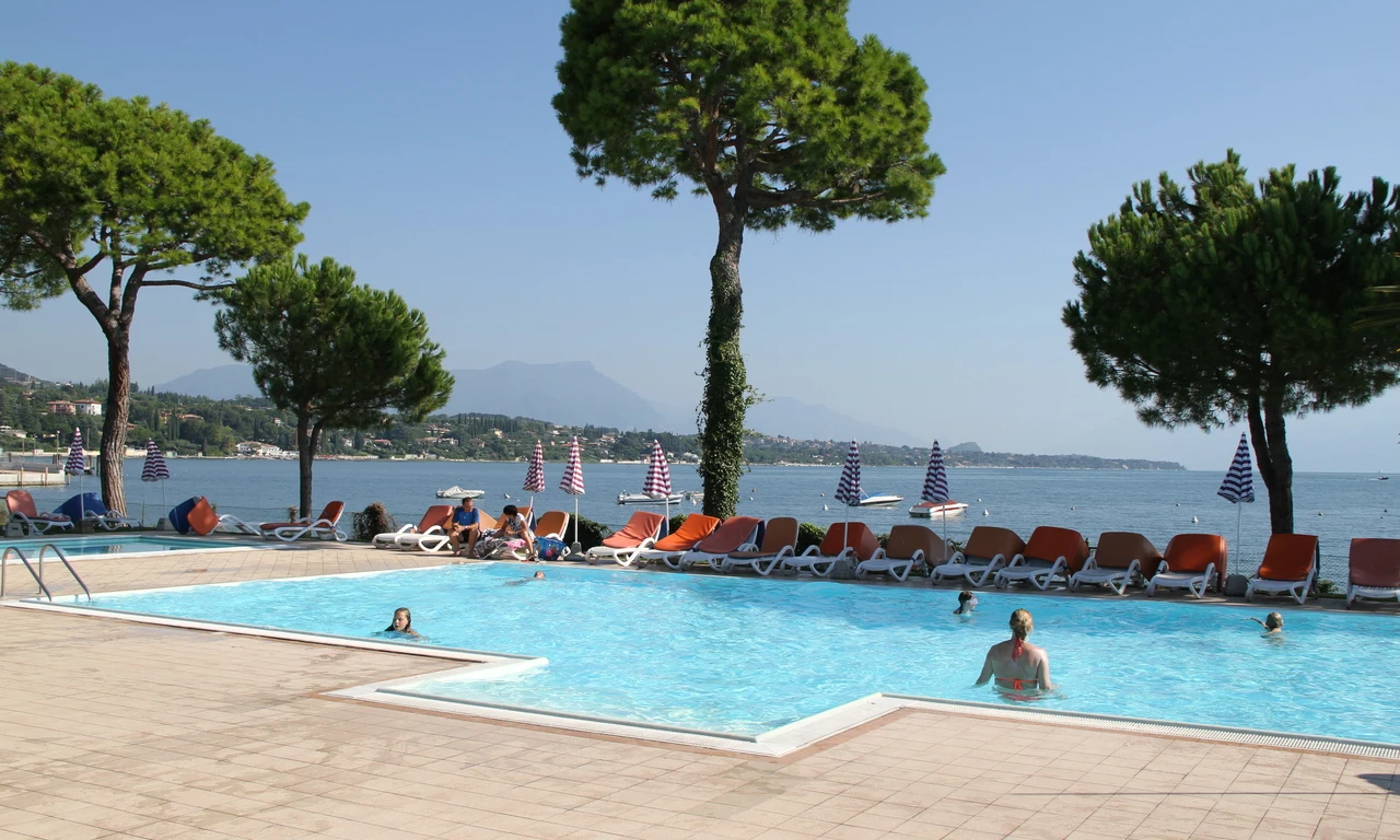 Pool with swimmers next to loungers and umbrellas, near a lake with trees and distant mountains at Front Lake Resort Le Corti del Lago