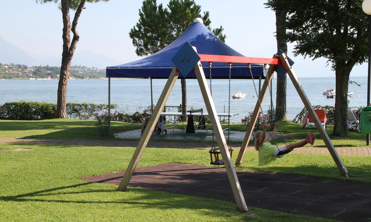 Child swinging on a playground swing set beside a blue tent, near a tranquil lake with boats at Front Lake Resort Le Corti del Lago