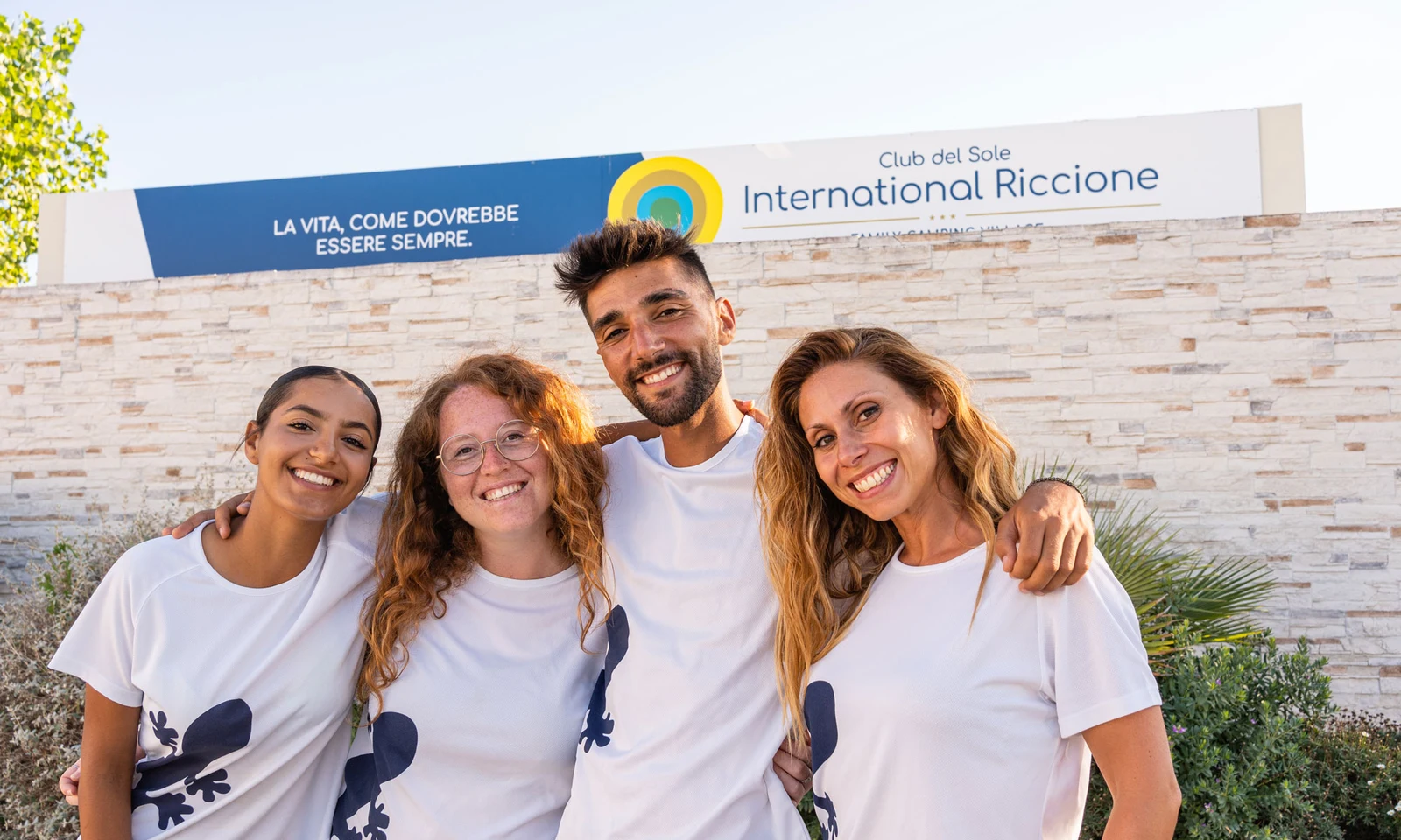 Four people smiling with arms around each other in front of a stone wall, outdoors at International Riccione Family Camping Village